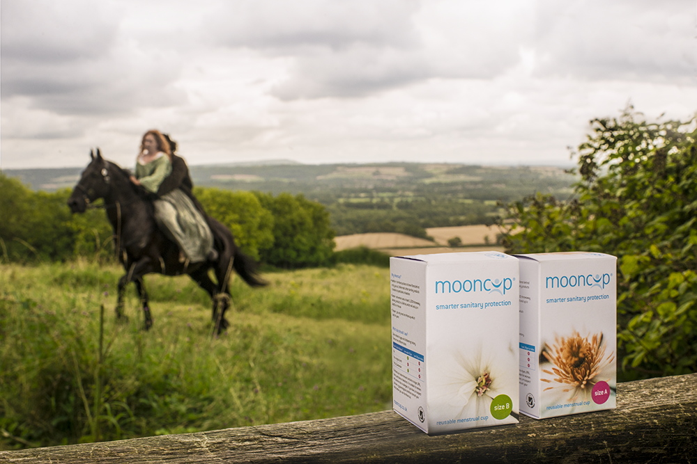 Period drama actors riding on a horse in a field with two Mooncup menstrual cups balancing on a fence in the foreground. 
