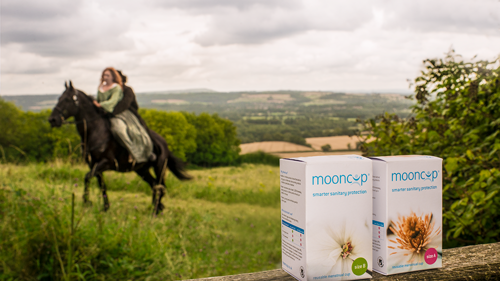 Period drama actors riding on a horse in a field with two Mooncup menstrual cups balancing on a fence in the foreground. 
