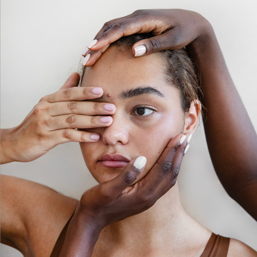 Woman's head and shoulers, she is covering her eye with her hand and someone else's hands are on the top of her head and around her cheek. 